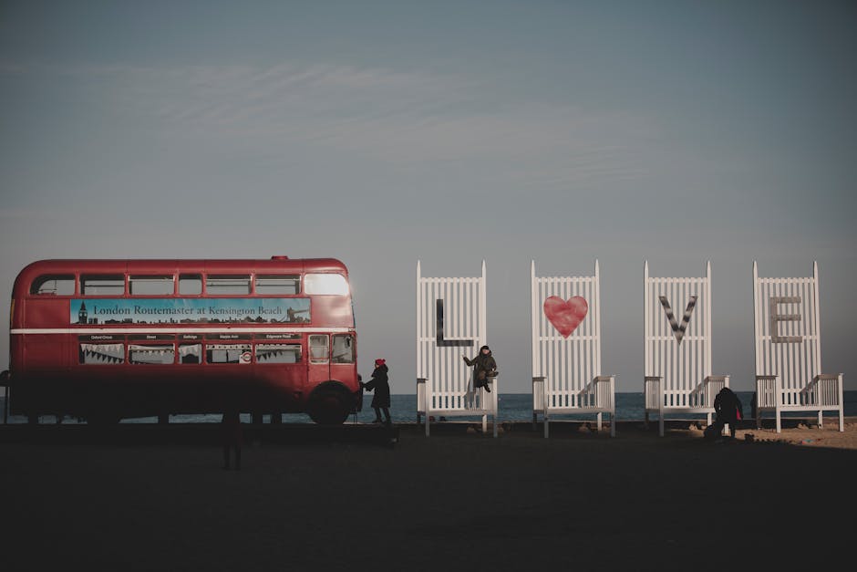 A red double-decker London Routemaster bus parked outdoors against a clear sky with some wispy clouds, featuring a large advertisement on its side promoting Kensingon Beach. The bus is stationary on a