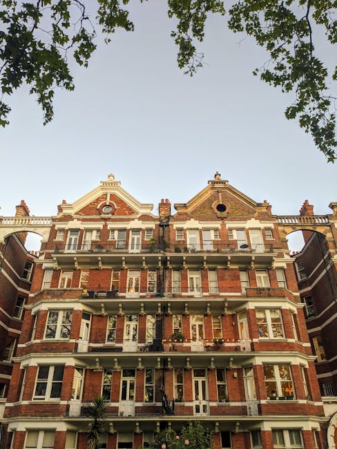 A long, narrow balcony or exterior corridor of a residential property with white-painted brick walls, supported by white columns and posts. The space is lined with planters containing dried or yellowi