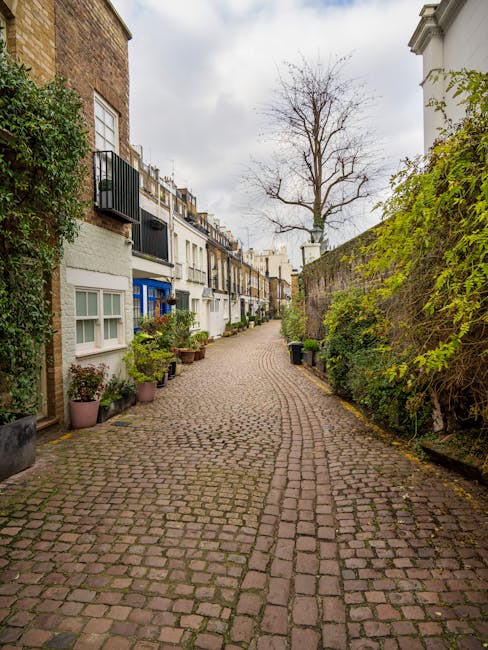 A narrow cobbled street in a residential area with multi-storey terraced houses on the left, each featuring white or cream facades, large windows, and small balconies with railings. Potted plants and 
