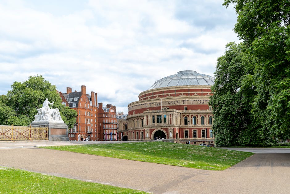 A wide-angle view of the Royal Albert Hall in South Kensington, showing its distinctive circular terracotta brick facade with large arched windows and ornate architectural details. In the foreground,