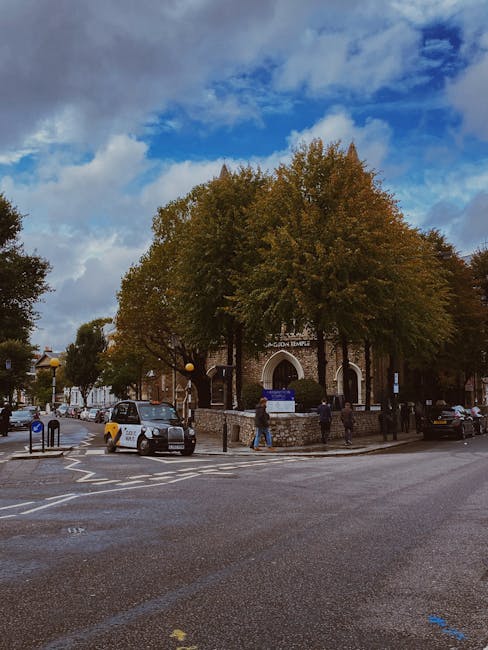 A photograph taken during daytime showing a street scene outside a chapel or church with a stone exterior, located on Gloucester Road in South Kensington. The building is surrounded by tall deciduous trees with green and brown foliage, indicating autumn. The sky above is partly cloudy with patches of blue sky visible. In front of the building, there are several parked cars, including a white van and a small grey car, on the paved street and roadside parking area. Pedestrians are walking on the sidewalk and crossing the street, while some appear to be carrying items or waiting. To the left, a tactile paving ramp and a pedestrian crossing are visible, along with street furniture like street lamps and bin stands. This scene contextualizes a typical urban area in South Kensington, often associated with house removals, furniture transport, or packing and moving activities by [COMPANY_NAME], supporting the relocation services for Gloucester Road flats.