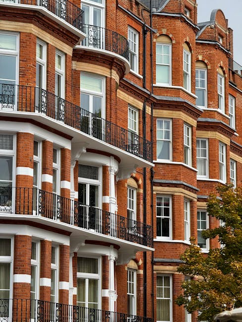 Exterior view of a multi-storey Victorian-style residential building with red brick walls, white decorative trim, and black wrought-iron balconies on each floor. The building features large sash windows, some with partially open curtains, set in a curved and angular facade. A small tree with green foliage is visible at the bottom right of the image, partially obscuring the lower part of the building. The photograph is taken during daylight with natural light illuminating the scene, highlighting the architectural details. The building appears suitable for home relocation or furniture transport services, such as those provided by South Kensington removals specialists, who may assist with packing, loading, and transporting household items from properties like this.