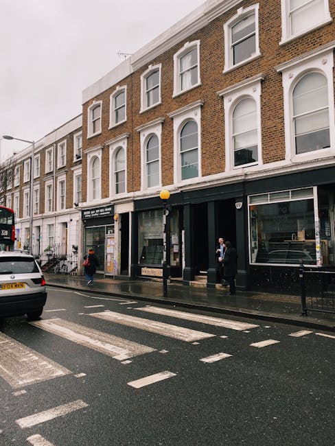 A photograph taken during daytime showing a street scene outside a chapel or church with a stone exterior, located on Gloucester Road in South Kensington. The building is surrounded by tall deciduous trees with green and brown foliage, indicating autumn. The sky above is partly cloudy with patches of blue sky visible. In front of the building, there are several parked cars, including a white van and a small grey car, on the paved street and roadside parking area. Pedestrians are walking on the sidewalk and crossing the street, while some appear to be carrying items or waiting. To the left, a tactile paving ramp and a pedestrian crossing are visible, along with street furniture like street lamps and bin stands. This scene contextualizes a typical urban area in South Kensington, often associated with house removals, furniture transport, or packing and moving activities by [COMPANY_NAME], supporting the relocation services for Gloucester Road flats.